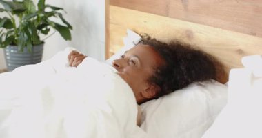 Kadın yatakta dinleniyor, beyaz battaniyeyle örtülmüş, evinde dinleniyor. Rahatlık, rahatlık, rahatlık, sükunet, huzur, rahatlık.