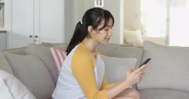 Telefon ekranına bakıyorum, Asyalı bir kadın açık gri koltukta güncellemeleri okumak için tıklıyor. Teknoloji, iletişim, rahatlık, rahatlık, minimalizm, yaşam tarzı, rahatlama