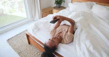 Yatakta dinlenen, akıllı telefon videosu izlerken gülümseyen bir kadın. Teknoloji, rahatlama, rahatlama, konfor, eğlence, yatak odası.