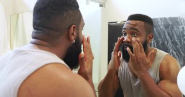 Afrika kökenli Amerikalı bir adam modern banyoya cilt bakım ürünleri uyguluyor, ayna yansıması üzerine çalışıyor. Tımar, yansıtma, cilt bakımı, banyo, modern sağlık, kişisel bakım.