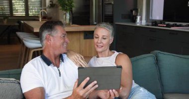 Evdeki kanepede oturan yaşlı çift, içerik için tablet yerleştiren adam, tıkırdayan kadın, gülen adam. Birlikte yaşama, samimiyet, rahatlama, modern, yaşam tarzı, teknoloji, konfor