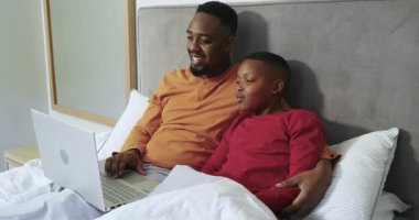 Afrika kökenli Amerikalı baba ve oğul pist taraması yapıyor. Yatakta bağlanmak için gümüş dizüstü bilgisayarla uğraşıyorlar. Aile, bağ, teknoloji, rahatlık, rahatlık, sıcaklık, ev