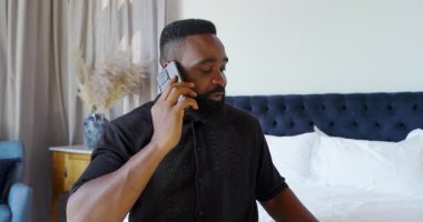 African American man sitting on neatly made bed using laptop at hotel room, focusing on work. Laptop, bed, hotel, casual, productivity, relaxation, work