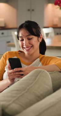 Dikey video: Asyalı kadın evde dinleniyor, kanepeye yaslanıyor, kameraya hafifçe gülümsüyor. Rahatlama, rahatlama, konfor, yaşam tarzı, sükunet, mutluluk.