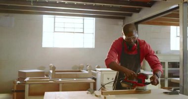 Afrika kökenli Amerikalı erkek marangoz zımpara makinesini alıyor, zımparalıyor ve tezgahta tahtayı bitiriyor. Zanaatkârlık, işçilik, marangozluk, atölye, elektrikli aletler, odaklanma, el işçiliği