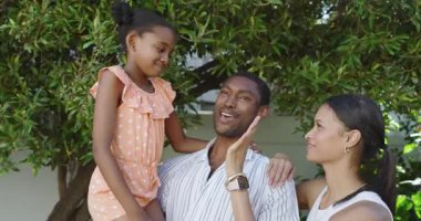 Afrikalı Amerikalı baba kızını tutarken anne yemyeşil ağacın altında gülümsüyor. Çeşitli aileler, ebeveynlik, bağlılık, açık hava, mutluluk, doğa