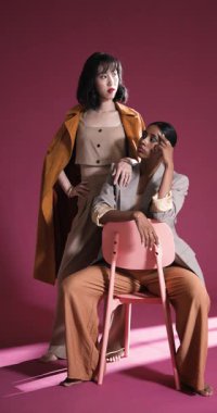 Dikey video: Modern stüdyoda pembe sandalyeyle poz veren şık giyinen kendine güvenen kadınlar. Moda, özgüven, tarz, zarafet, şıklık.