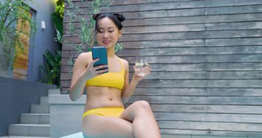 Akıllı telefonu kaydıran kadın, camdan su yudumlayan ve arka bahçedeki havuz kenarına ayak basan kadın. Rahatlama, rahatlama, açık hava, sıcak, güneşli, yaşam tarzı, moda.