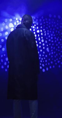 Dikey video: Sanatsal stüdyo ortamında mavi ışıkların altında duran koyu renk ceketli adam. Moda, portre, yaratıcı, ışıklandırma, erkek, ceket