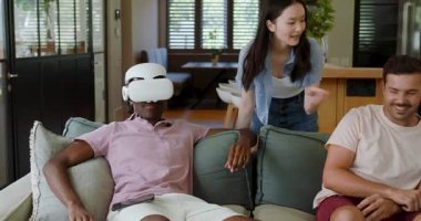 Çeşitli arkadaşlar, oturma odasında tepki veren Afro-Amerikalı bir adama VR kulaklık takan kadını izliyorlar. Günlük, sosyal, modern, yaşam tarzı, teknoloji, eğlence, rahatlama