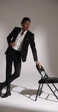 Dikey video: Siyah takım elbiseli model stüdyoda sandalyeyle güven içinde poz veriyor. poz, zarafet, özgüven, tarz