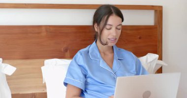 Mavi pijamalı bir kadın yatakta dizüstü bilgisayar kullanıyor. Düşünceli bir şekilde pencereden dışarı bakıyor. Rahatlama, rahatlık, sükunet, teknoloji, tefekkür, gece vakti