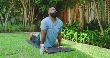 Siyah yoga minderinde yoga yapan Afrikalı Amerikalı bir adam yeşil çimlerin üzerinde, su şişesi de yanında. Yoga, meditasyon, sağlık, sükunet, açık hava, sağlık, farkındalık