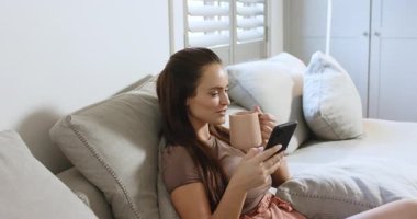 Kanepeye kurulmak, yirmili yaşlardaki bir kadın akıllı telefonu kaydırmak ve rahatlamak için fincanı yudumlamak, boşluğu kopyalamak. Rahatlık, rahatlık, ev, huzur, rahat, rahat, farkındalık.