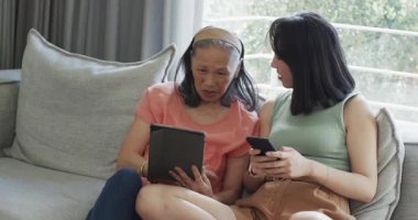 Çinli anne ve kız, gri kanepeye telefon yükleyip tablet karıştırarak resim paylaşıyorlar. Nesiller arası, bağlantı, teknoloji, aile, bağ, öğrenme, iletişim