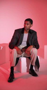 Dikey video: Moda modeli şık bir takım elbise içinde küp üzerinde oturuyor, stüdyoda düşünceli görünüyor. çağdaş, zarif, portre, profesyonel