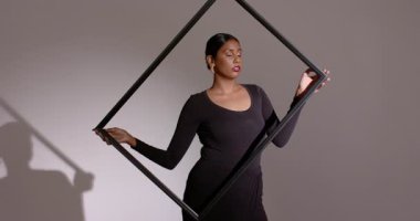 Belden siyah bir çerçeve kaldırıyor, Hintli bir kadın çerçeveyi döndürüyor, yüzünü gri stüdyo duvarına yaslıyor. Çağdaş, minimalist, sanatsal, moda, siluet, dinamik, zarafet