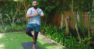 Afro-Amerikalı bir adam açık havada yoga yapıyor, yemyeşil bir ortamda, sıkılmış ellerle odaklanıyor. Meditasyon, sükunet, doğa, fitness, farkındalık, sağlık, yeşillik