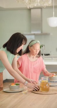 Dikey video: Mutfak masasında kızıyla konuşurken yaşlı bir kadın meyve suyu dolduruyor. Aile, kaynaşma, sohbet, misafirperverlik, yaşam tarzı, içecek