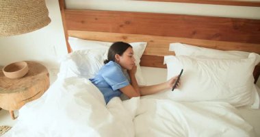 Yatakta elinde akıllı telefon tutarak gülen bir kadın, evde sabah rahatlığının tadını çıkarıyor. Yaşam tarzı, teknoloji, eğlence, mutluluk, sabah rutini, bağlantı