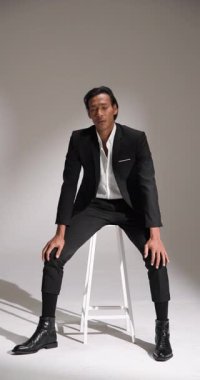 Dikey video: kendine güvenen şık takım elbiseli adam stüdyoda beyaz sandalyeyle poz veriyor. Moda, tarz, zarafet, sofistike, erkek model, resmi kıyafet.