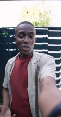 Dikey video: Güneş ışığı, kameralı Afro-Amerikan bir adamın vlog için avluya girmesini teşvik ediyor. Vlog, yaşam tarzı, açık hava, yeşillik, gündelik, doğa, dikey video