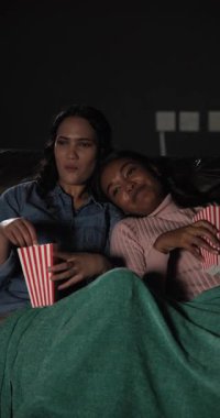 Vertical video: Suspense sparking Diverse female friends sharing popcorn on sofa flinching. Friendship, companionship, relaxation, entertainment, cozy, casual, vertical video