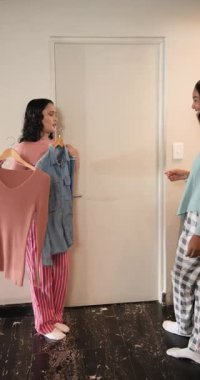 Dikey video: Askılara bakan kadın arkadaşlar ve evde süveter ve tişört giyme arasında seçim yapmak. Arkadaşlık, moda, gündelik, yaşam tarzı, ev, etkileşim, dikey video