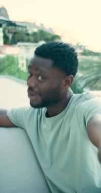 Dikey video: Afro-Amerikalı adam nefes veriyor sonra telefonla konuşuyor ve balkonda selamlaşmak için el sallıyor. Açık hava oturma, günlük yaşam tarzı, canlı çevre, modern mimari, rahat ruh hali, gençlik enerjisi, dikey video