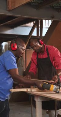 Vertical video: After rubbing plank African American team cutting and sanding wood at shop bench. Craftsmanship, teamwork, industrial, rustic, manual labor, construction, vertical video