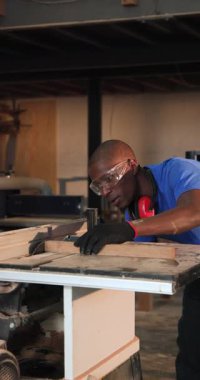 Dikey video: Afrika kökenli Amerikalı orta yaşlı bir adamın montaj için tezgah kestiğini duymak. Zanaatkar, atölye, endüstriyel, marangozluk, kesinlik, güvenlik, dikey video