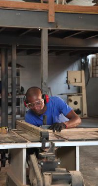 Dikey video: Afrikalı Amerikalı bir adam atölyedeki kalasları keserek inceliyor. İşçilik, marangozluk, endüstriyel, el işi, odaklanma, güvenlik, dikey video