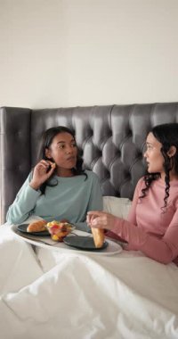 Dikey video: Kahvaltı tepsisi alındıktan sonra çeşitli kadın arkadaşlar yatakta pasta ve meyve yiyorlar. Arkadaşlık, brunch, eğlence, yaşam tarzı, rahat, samimi, dikey video