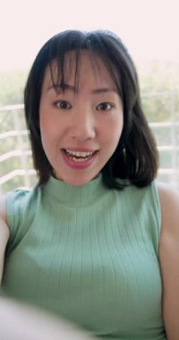 Dikey video: Kameraya bakan, Çinli bir kadın, balkon kanepesinde saçlarını düzeltirken konuşuyor. Açık hava, yaşam tarzı, gündelik, yeşillik, doğal ışık, iletişim, dikey video