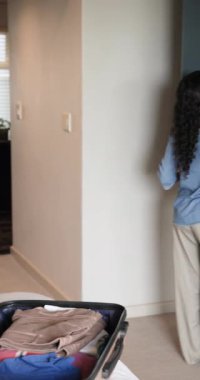 Vertical video: Diverse female friends opening suitcase and hanging clothes with hangers in bedroom. Friendship, packing, travel, lifestyle, casual, domestic, vertical video