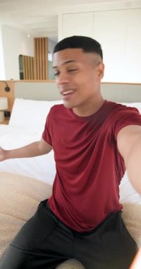 Dikey video: Dinleme kaydı, erkek kameraya el sallıyor, konuşuyor, gülüyor, yatak odasında telefonu eğiyor. Rahatlama, iletişim, günlük, yaşam tarzı, modern, samimi, dikey video