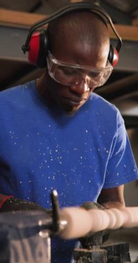 Dikey video: Dükkandaki torna tezgahına oyma oymacılığı yapan Afro-Amerikalı bir adam, fotokopi alanı. Zanaatkar, atölye, marangoz, marangozluk, dikey video