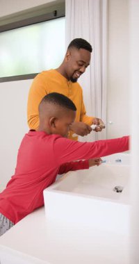 Dikey video: Afrikalı Amerikalı baba ve oğlu banyo lavabosunda diş fırçalayıp macun kullanıyor. Aile, bağlanma, hijyen, bakım, rutinler, ev içi, dikey video