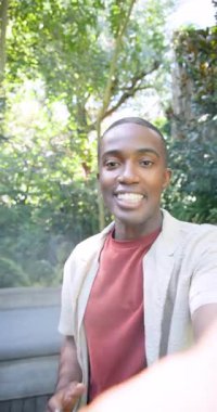 Dikey video: Bahçe bankında görüntüsünü düzelten, gülümseyen Afrikalı Amerikalı bir adam tutmak. Vlog, outdoor, yemyeşil, doğal, yaşam tarzı, samimi, dikey video