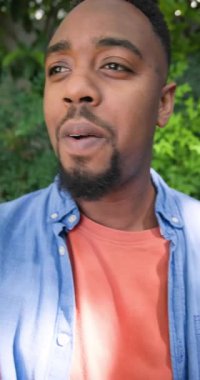 Dikey video: kot gömlekli Afro-Amerikalı adam bahçede kameraya yansıyan görüntüyü paylaşıyor. Portre, doğa, açık hava, günlük, sağlık, iletişim, dikey video