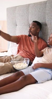 Dikey video: Uzaktan kumandalı Afro-Amerikan baba ve oğlu yatakta patlamış mısır yiyerek televizyonu işaret ediyor. Aile, eğlence, eğlence, konforlu, sevecen, dikey video