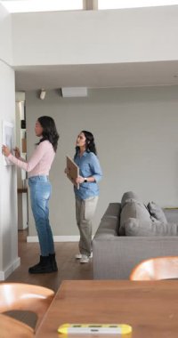 Dikey video: Kadın arkadaşlar eve resim getiriyor, katları kullanarak duvara asıyorlar, sarılıyorlar. Ev dekorasyonu, iç tasarım, arkadaşlık, işbirlikçi, modern, yaşam tarzı, dikey video