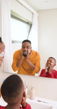 Dikey video: Afro-Amerikan baba oğluna banyo lavabosunda köpük köpürterek tıraş olmayı öğretiyor. Aile bağı, bakım, cilt bakımı, hijyen, eğitim, ebeveynlik, dikey video