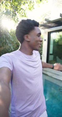 Dikey video: Afro-Amerikan adam elinde akıllı telefon, arka bahçede havuz kenarında oturma odası tutuyor. Açık hava, yaşam tarzı, minimalist, boş zaman, mimari, rahatlama, dikey video