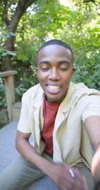 Dikey video: Afrika kökenli Amerikalı erkek güvertede bankta kamerayla konuşuyor, lensleri koruyor. Doğa, açık hava, video, yaşam tarzı, dinginlik, iletişim, dikey video