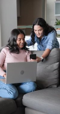 Dikey video: Kanepede oturan, dizüstü bilgisayarı olan ve telefona bakıp kahveyi yudumlayan kadın arkadaşlar. İşbirliği, arkadaşlık, eğlence, ev, modern yaşam tarzı, dikey video