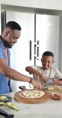 Dikey video: Afro-Amerikalı baba kabuğu yerleştiriyor ve oğlu pizza hazırlamak için peynir rendeliyor. Aile, aşçılık, el yapımı, sıcak, kaynaştırıcı, öğretici, dikey video