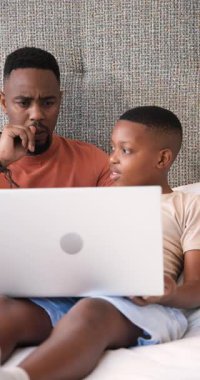 Dikey video: Kanepede dizüstü bilgisayar izleyen Afrikalı baba ve oğul, erkek işaret eden, baba kafa sallayan. Aile, bağ, eğitim, teknoloji, eğlence, iletişim, dikey video