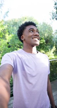 Dikey video: Tahta güvertede akıllı telefonu genişletmek, Afro-Amerikalı bir adam izleyiciyi el sallayarak selamlıyor. Açık hava, boş zaman, yaşam tarzı, gündelik, doğa, iletişim, dikey video