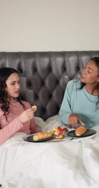 Vertical video: Two women sitting up eating breakfast in bed with tray of croissants and fruit bowl. Intimacy, companionship, leisure, relaxation, warm, tender, vertical video
